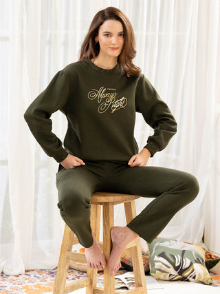 Woman wearing a green sweatshirt and pants sitting on a stool with white curtains in the background